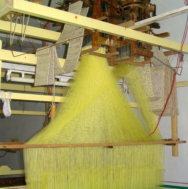 The traditional loom for making Kanchipuram sari. Those white cards with holes in it defines the patterns/designs in the Sari. Also seen is the yellow silk thread that fanning out from the top of the loom.